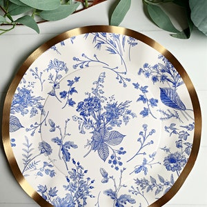 May include: A white paper charger plate with a blue floral pattern and a gold rim. The plate features a variety of blue flowers and leaves on a white background.