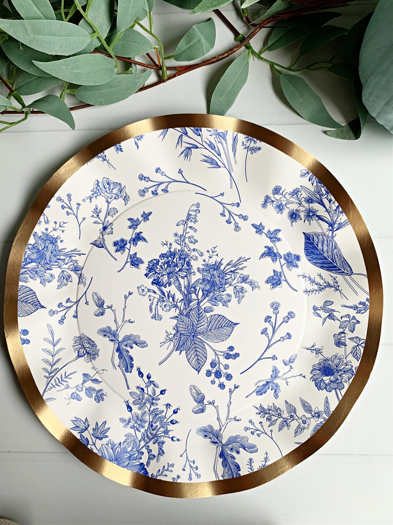 May include: A white paper charger plate with a blue floral pattern and a gold rim. The plate features a variety of blue flowers and leaves on a white background.