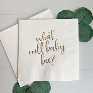 May include: White paper napkin with gold lettering that reads "what will baby bee?"