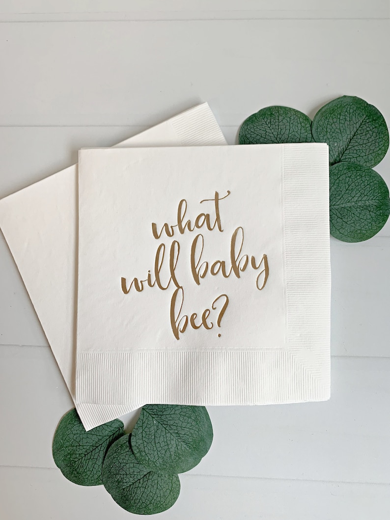 May include: White paper napkin with gold lettering that reads "what will baby bee?"