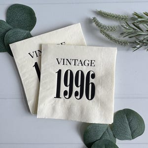 May include: Two off-white paper napkins with the word "VINTAGE" and the number "1996" printed in black. The napkins are surrounded by green leaves and sprigs on a white wooden surface.