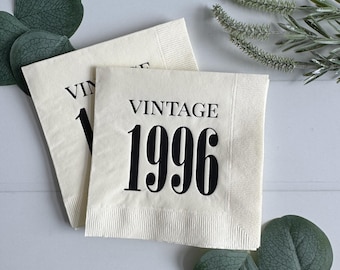 May include: Two off-white paper napkins with the word "VINTAGE" and the number "1996" printed in black. The napkins are surrounded by green leaves and sprigs on a white wooden surface.