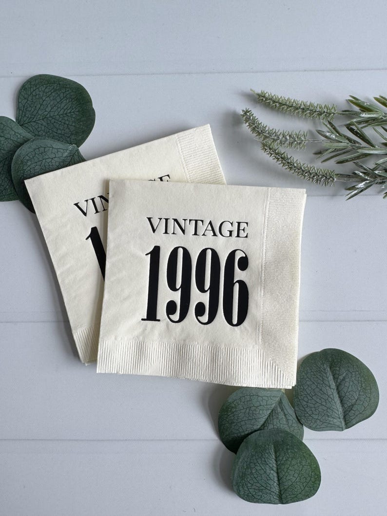 May include: Two off-white paper napkins with the word "VINTAGE" and the number "1996" printed in black. The napkins are surrounded by green leaves and sprigs on a white wooden surface.