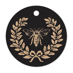 May include: A black circle with a gold bee and laurel wreath design. The bee is in the center of the wreath and has a detailed design. The wreath is made of leaves and berries.