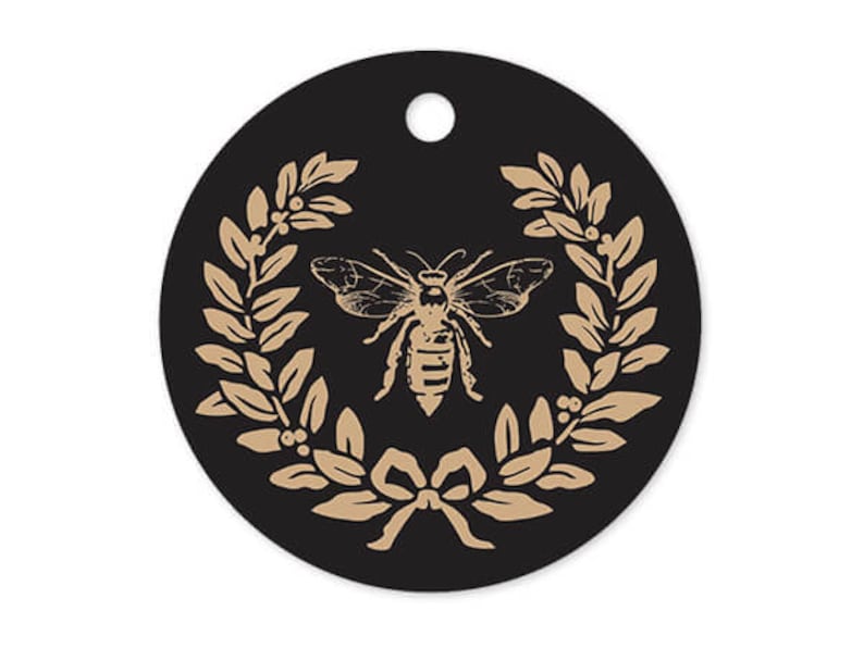May include: A black circle with a gold bee and laurel wreath design. The bee is in the center of the wreath and has a detailed design. The wreath is made of leaves and berries.