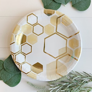May include: White paper party plates with a gold honeycomb pattern. The plates are surrounded by green eucalyptus leaves.