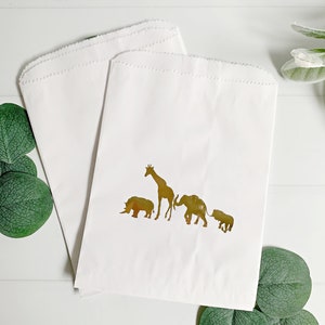 May include: Two white paper bags with gold foil animal silhouettes. The animals are a giraffe, rhinoceros, elephant, and a smaller elephant. The bags are perfect for party favors or gifts.
