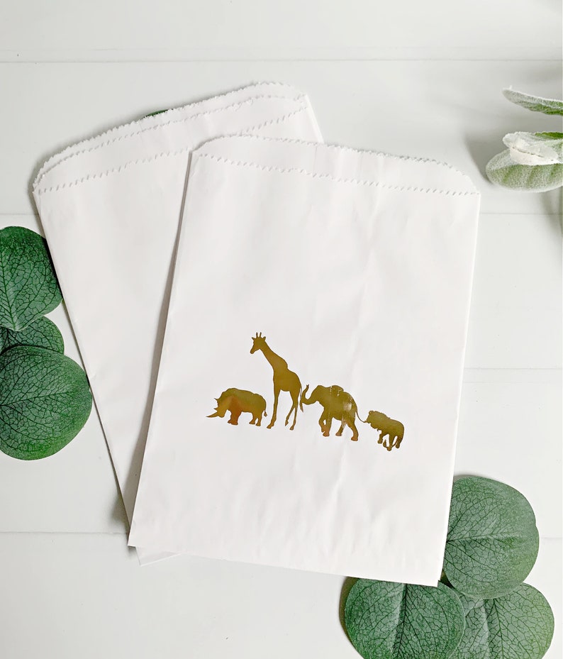May include: Two white paper bags with gold foil animal silhouettes. The animals are a giraffe, rhinoceros, elephant, and a smaller elephant. The bags are perfect for party favors or gifts.