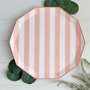 May include: Pink and white striped paper party plate with a gold rim. The plate is on a white wooden surface with green leaves around it.