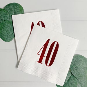 May include: Two white paper napkins with a red number 40 printed on them. The napkins are folded and placed on a white surface with green leaves.