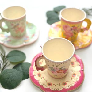 May include: Three paper teacups and saucers with floral patterns. The cups have handles and are decorated with roses and other flowers. The saucers have scalloped edges and a pink border. The background is white with green leaves.