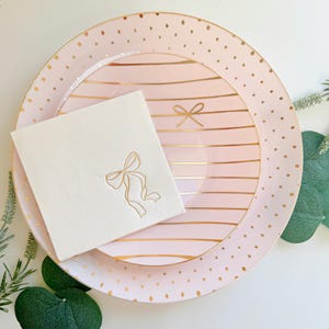 May include: A set of pink and gold decorative plates and a napkin. The large plate has a pink background with gold polka dots. The smaller plate has a pink background with gold stripes. The napkin is white with a gold bow design.
