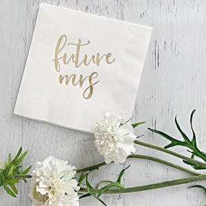 Puede incluir: Servilleta de papel blanca con letras doradas que dicen "future mrs". La servilleta est&aacute; sobre una superficie de madera blanca con flores blancas.