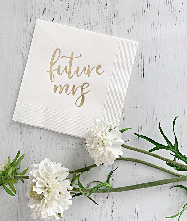 Puede incluir: Servilleta de papel blanca con letras doradas que dicen "future mrs". La servilleta est&aacute; sobre una superficie de madera blanca con flores blancas.