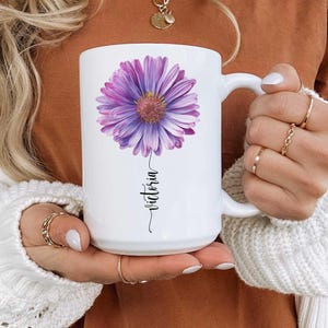 May include: White ceramic mug featuring a large purple and yellow flower design. The name "Victoria" is written in a cursive font below the flower. The mug has a standard handle and is held by a person.