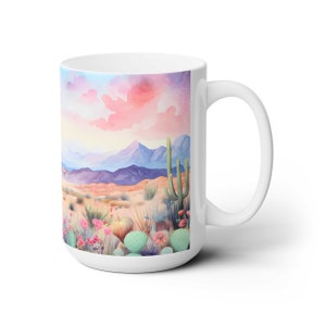 May include: White ceramic mug with a colorful watercolor illustration of a desert landscape with cacti and flowers. The background features a pink and purple sunset sky with mountains in the distance.