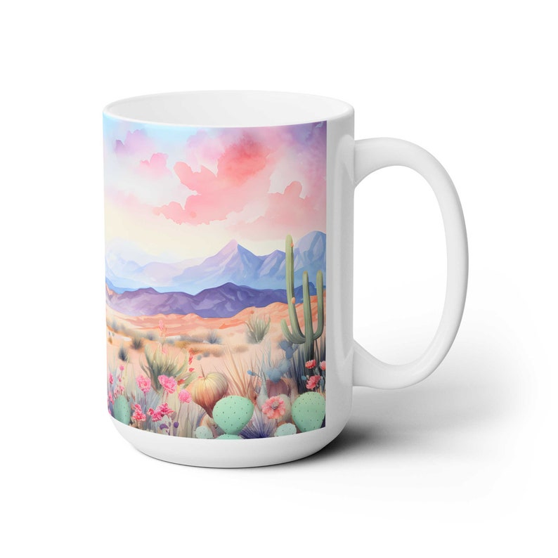 May include: White ceramic mug with a colorful watercolor illustration of a desert landscape with cacti and flowers. The background features a pink and purple sunset sky with mountains in the distance.