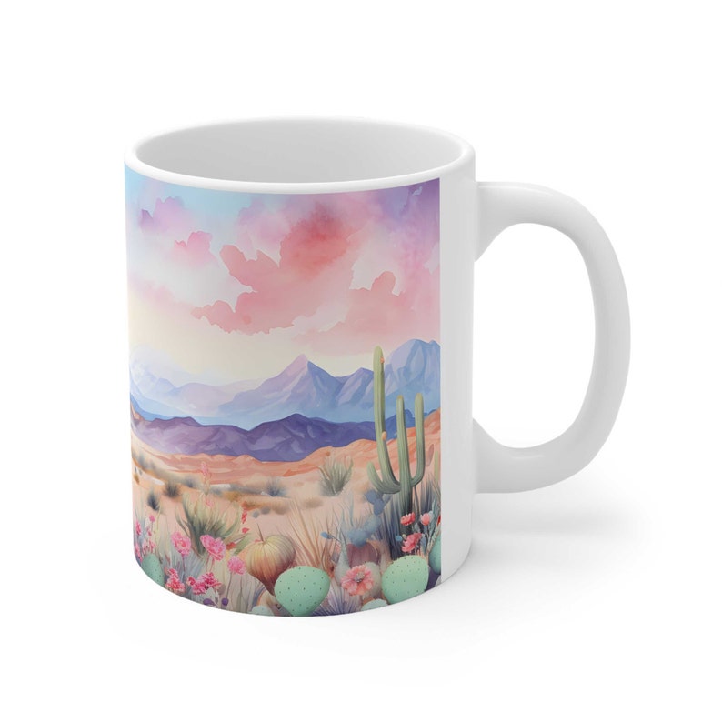 May include: White ceramic mug with a watercolor illustration of a desert landscape. The scene features a pink and purple sunset over a mountain range with cacti and prickly pear plants in the foreground.