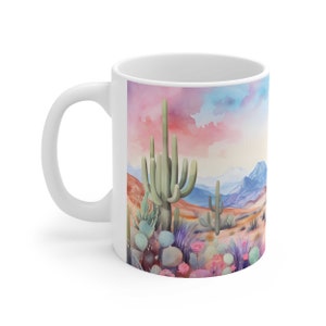May include: White ceramic mug with a colorful watercolor illustration of a desert landscape featuring tall cacti, prickly pear cacti, and pink flowers against a backdrop of mountains and a pink and blue sky.