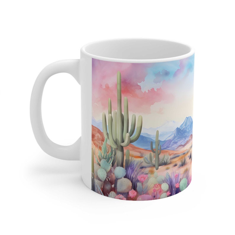 May include: White ceramic mug with a colorful watercolor illustration of a desert landscape featuring tall cacti, prickly pear cacti, and pink flowers against a backdrop of mountains and a pink and blue sky.