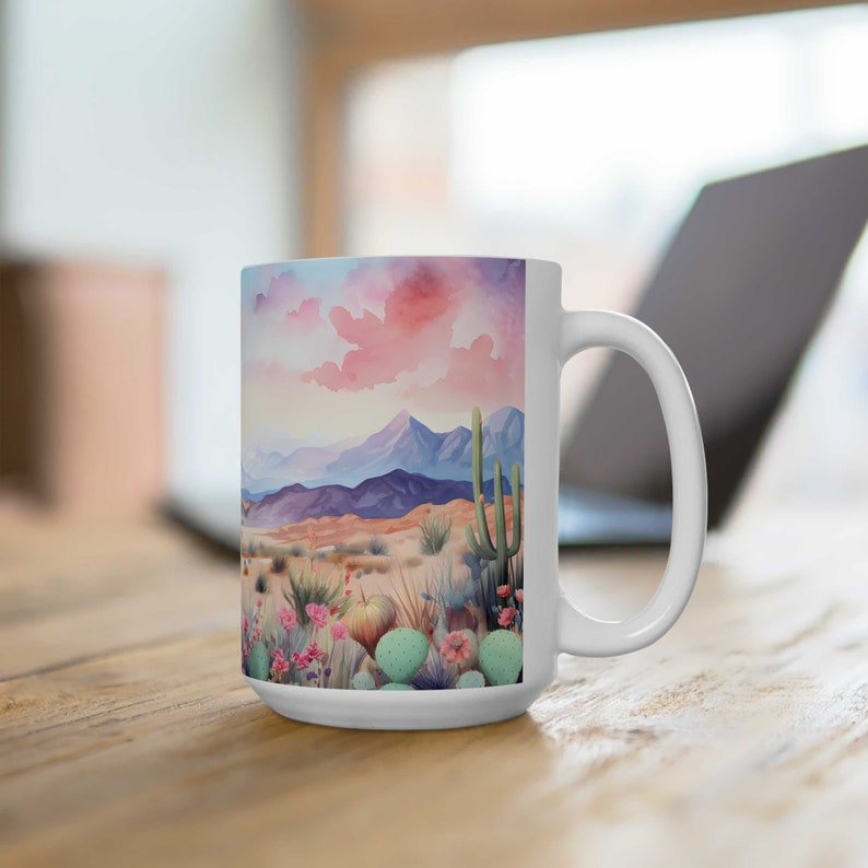 May include: White ceramic mug with a watercolor illustration of a desert landscape with mountains, cacti, and pink clouds.