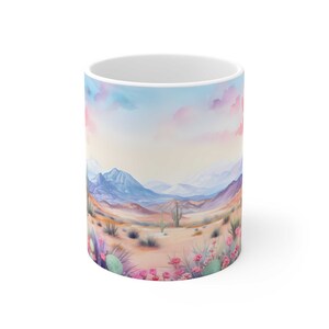 May include: A white ceramic mug with a watercolor design of a desert landscape. The scene features mountains, cacti, and pink flowers.