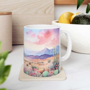 May include: White ceramic mug with a colorful watercolor illustration of a desert landscape with mountains, cacti, and flowers. The mug has a handle and is sitting on a beige coaster.