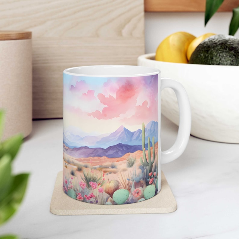 May include: White ceramic mug with a colorful watercolor illustration of a desert landscape with mountains, cacti, and flowers. The mug has a handle and is sitting on a beige coaster.