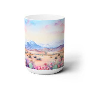 May include: White ceramic mug with a watercolor design of a desert landscape. The scene features mountains, cacti, and pink flowers in the foreground. The sky is a soft blue with pink clouds.