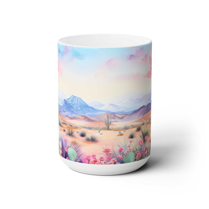 May include: White ceramic mug with a watercolor design of a desert landscape. The scene features mountains, cacti, and pink flowers in the foreground. The sky is a soft blue with pink clouds.