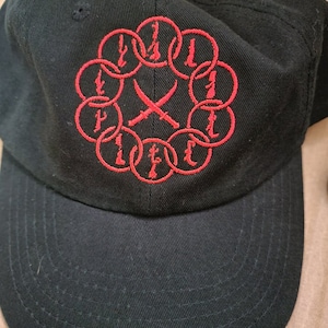 May include: Black baseball cap with a red embroidered design of a circle with swords crossed in the center and the letter "L" repeated around the circle.
