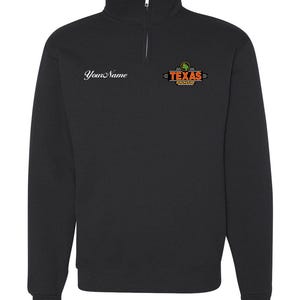 Puede incluir: Sudadera negra con cremallera de cuarto de cuello. En el pecho izquierdo, el texto "Your Name" en cursiva. En el pecho derecho, un logotipo con las palabras "TEXAS ROADHOUSE" en naranja y verde.