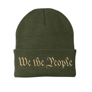 Puede incluir: Gorro verde oliva con el texto "We the People" en una fuente decorativa. El gorro es de estilo con borde doblado, hecho de material de punto. El texto es de color beige.