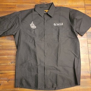 Puede incluir: Camisa negra de manga corta con dos bolsillos en el pecho. La camisa presenta la palabra "TOOL" en letras blancas en el bolsillo derecho y un diseño de llama en el bolsillo izquierdo. La camisa está hecha de una tela duradera.