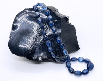 May include: A necklace of oval, translucent blue beads rests on a dark, textured black stone. The beads are strung together, forming a loop. The background is plain white.