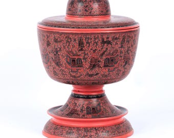 May include: A red and black lidded bowl on a pedestal base. The exterior surface is decorated with intricate black patterns featuring small figures, structures, and foliage. The bowl has a matching lid with a central knob and a wide, flared foot.