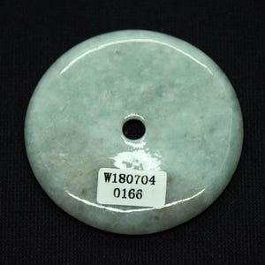 May include: A pale green jade pendant with a central hole. The circular stone has a smooth, polished surface and a subtle, natural color variation. A small white sticker with the numbers "W180704" and "0166" is affixed to the pendant.
