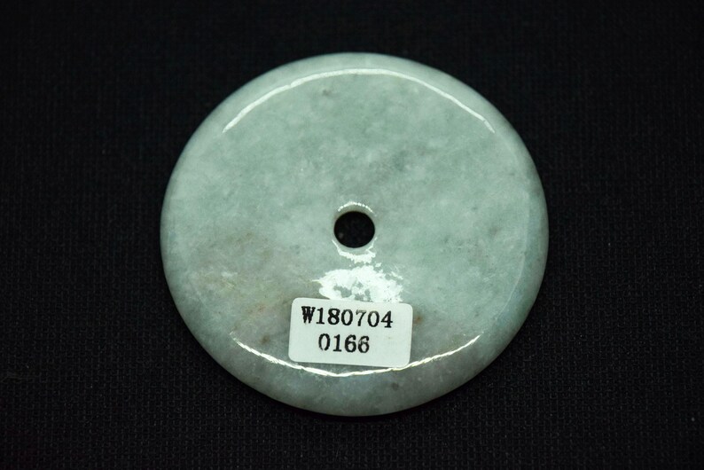 May include: A pale green jade pendant with a central hole. The circular stone has a smooth, polished surface and a subtle, natural color variation. A small white sticker with the numbers "W180704" and "0166" is affixed to the pendant.