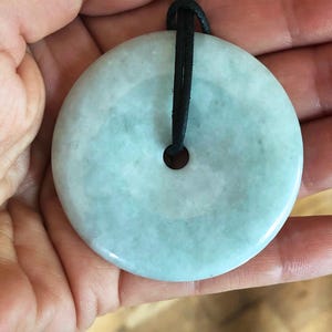 May include: A round, pale green jade pendant with a black cord. The pendant has a central hole and a smooth, polished surface. The jade has a subtle, translucent quality, and the cord is knotted at the top.