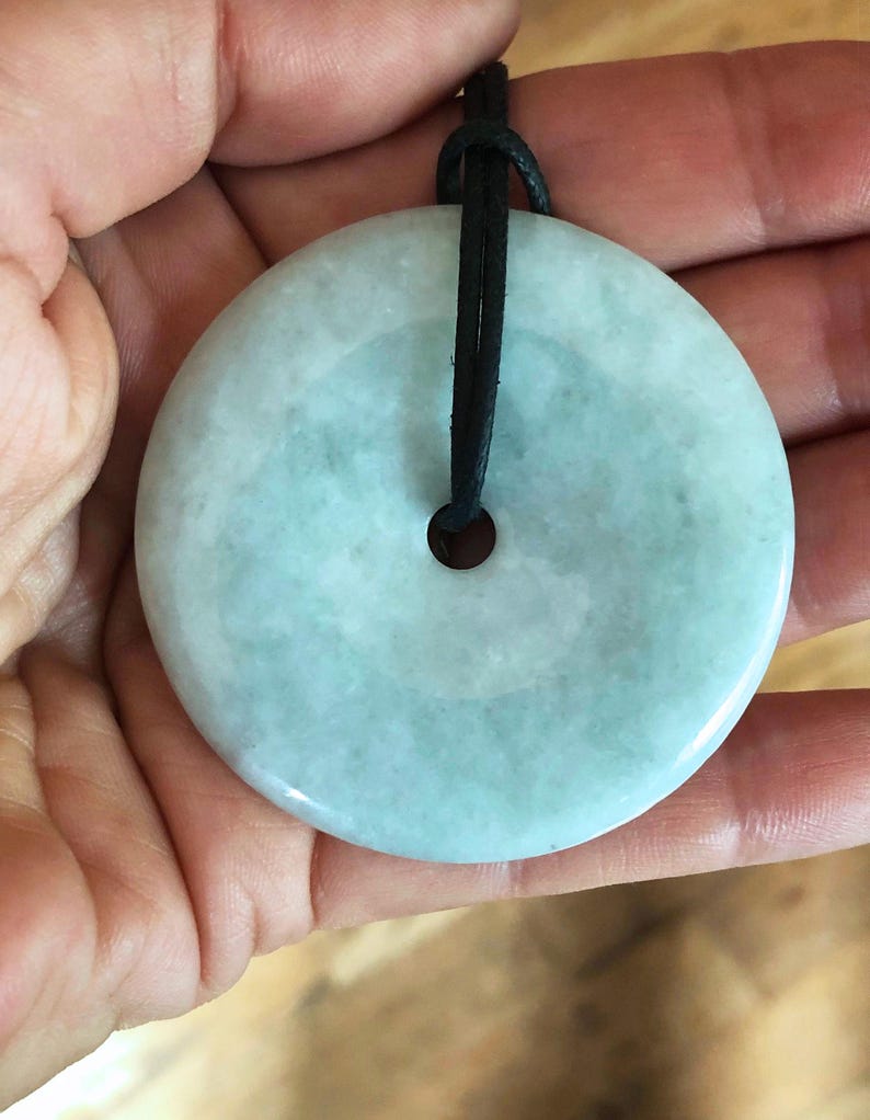 May include: A round, pale green jade pendant with a black cord. The pendant has a central hole and a smooth, polished surface. The jade has a subtle, translucent quality, and the cord is knotted at the top.