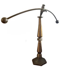 May include: A bronze-toned metal balancing toy featuring a central vertical stand with a hexagonal base. A curved horizontal arm is balanced at the top, with a small bird figure at the center and two spherical weights attached to each end of the arm.