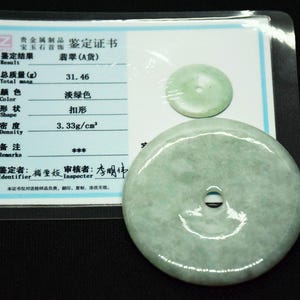 May include: A pale green jade pendant with a central hole, next to a smaller jade disc. A certificate in Chinese characters is visible in the background, encased in plastic.