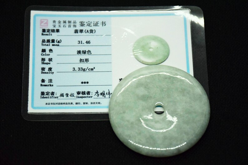May include: A pale green jade pendant with a central hole, next to a smaller jade disc. A certificate in Chinese characters is visible in the background, encased in plastic.