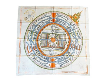 May include: A white silk scarf with a colorful map-like design. The map features a circular design with a central orange circle surrounded by blue, green, and yellow lines. The scarf has a white border with a small design in the corner.