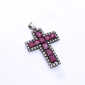 Può includere: Un pendente in argento a forma di croce, ornato con pietre preziose rosse ovali e pietre più piccole e trasparenti. La croce ha un'asola per attaccare una catena. Le pietre sono incastonate in argento.