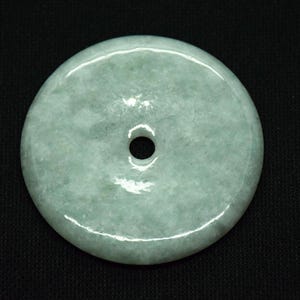 May include: A round, pale green jade pendant with a central hole. The stone has a smooth, polished surface with subtle variations in color. The pendant is set against a black background.