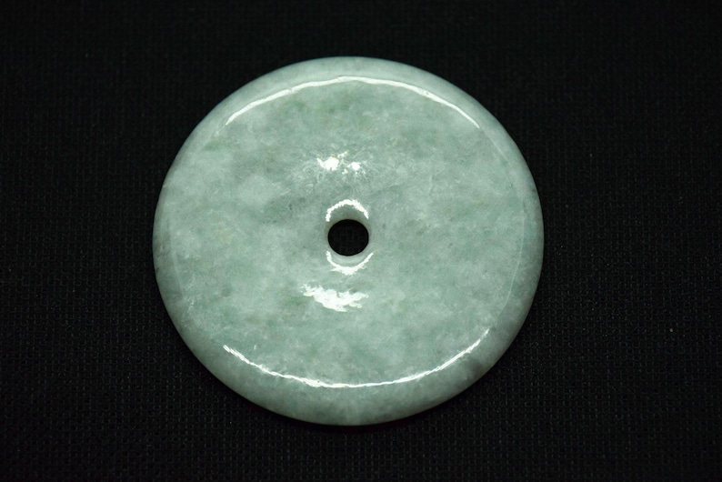 May include: A round, pale green jade pendant with a central hole. The stone has a smooth, polished surface with subtle variations in color. The pendant is set against a black background.