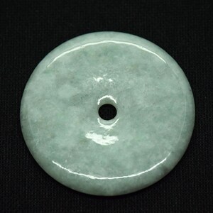 May include: A round, pale green jade pendant with a central hole. The stone has a smooth, polished surface with subtle variations in color. The pendant is set against a black background.