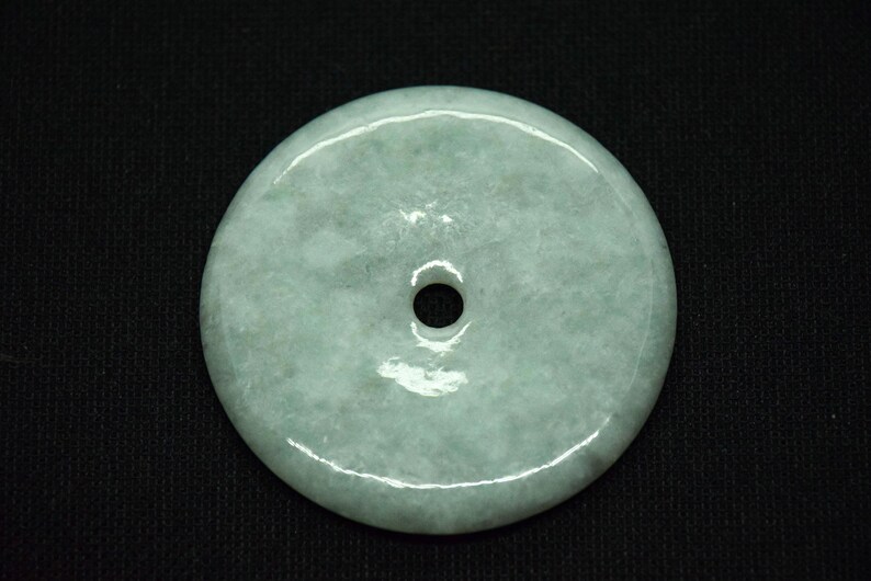 May include: A round, pale green jade pendant with a central hole. The stone has a smooth, polished surface with subtle variations in color. The pendant is set against a black background.