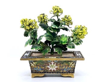 May include: A decorative metal sculpture of a chrysanthemum plant with yellow flowers and green leaves, set in a rectangular cloisonné planter. The planter features intricate floral patterns in blue, green, white, and gold enamel on a brass base.
